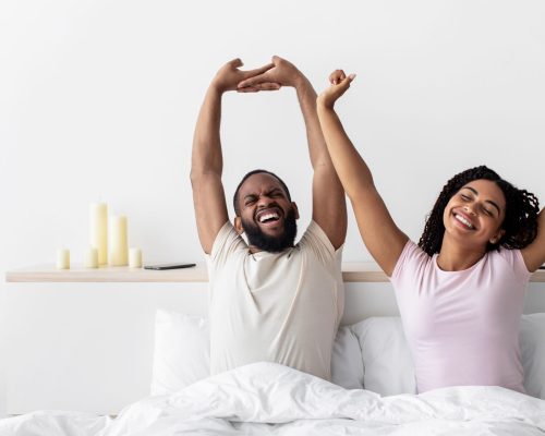 Glad happy young african american lady and guy woke up stretching body and enjoy morning on comfortable bed in bedroom, empty space. Rest together at weekend, vacation and free time at home, panorama